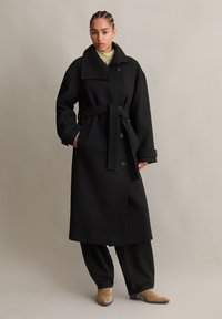Zwarte wollen trenchcoat met een brede kraag, een ceintuur in de taille en grote knopen. Model draagt donkere broeken en beige enkellaarsjes. Neutrale achtergrond.
