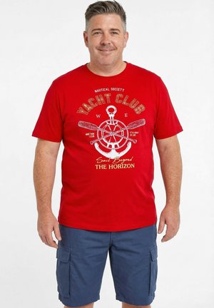 Uomo che indossa una t-shirt rossa con grafica "Yacht Club" e pantaloncini cargo blu, in piedi davanti a uno sfondo bianco semplice.