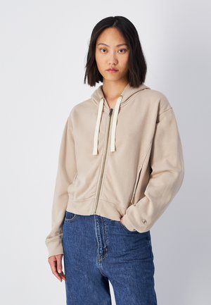 Sweat à zip beige avec capuche à cordon, coupe décontractée et texture douce. Comprend des poches avant et des poignets côtelés. Associé à un jean bleu.