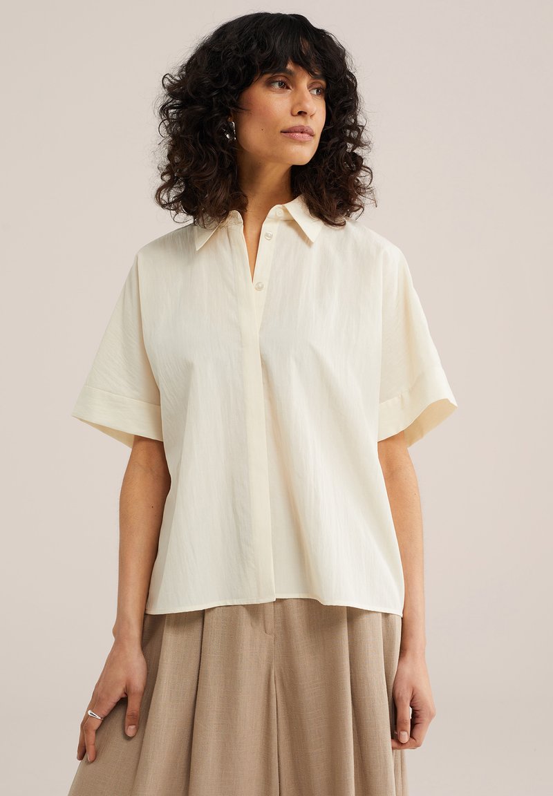 Femme aux cheveux bouclés foncés portant un chemisier ample à manches courtes crème et un pantalon large beige, debout devant un fond uni.