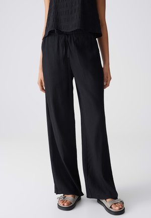 Femme portant un pantalon noir ample à cordon de serrage et un haut noir sans manches, debout avec des sandales grises à boucle sur un fond uni.