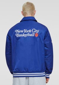 Niebieska nylonowa kurtka z białym napisem "New York City Basketball" i grafiką piłki z tyłu; paski na mankietach i dolnej krawędzi dodają szczegółu.