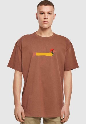 Brun oversized t-shirt med et cartoon billede af en dachshund, med en gul krop, rød hat og grøn tørklæde på forsiden.
