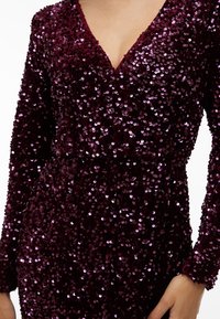 Nærbilde av en person som har på seg en dyp lilla paljettkjole med lange ermer og V-hals, som viser en del av hånden og brystet.