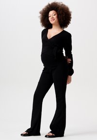 Schwarzer Umstandsoverall im Wickelstil mit langen Ärmeln, seitlichem Bindegürtel und weit ausgestellten Beinen, aus einem strukturierten Stoff gefertigt. Sandalen ergänzen das Outfit.