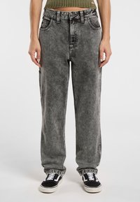 Pantalones de mezclilla gris tratado con ácido, de corte holgado, cintura alta y cinco bolsillos. Costuras negras y puños enrollados. Combinados con zapatillas negras.
