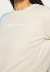 Žena na sobě má krémovou mikinu Calvin Klein Jeans s logem umístěným přes hrudník a světle modrým páskem přes rameno.