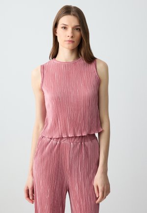 Jonge vrouw in een mouwloos roze geribd topje en bijpassende high-waisted broek, staand voor een effen lichtgrijze achtergrond.