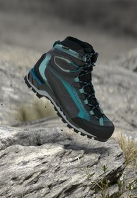 Μπλε και μαύρο πεζοπορικό μποτάκι με ανθεκτική σόλα που αιωρείται πάνω από βραχώδες έδαφος με αραιό γρασίδι και μικρά φυτά.