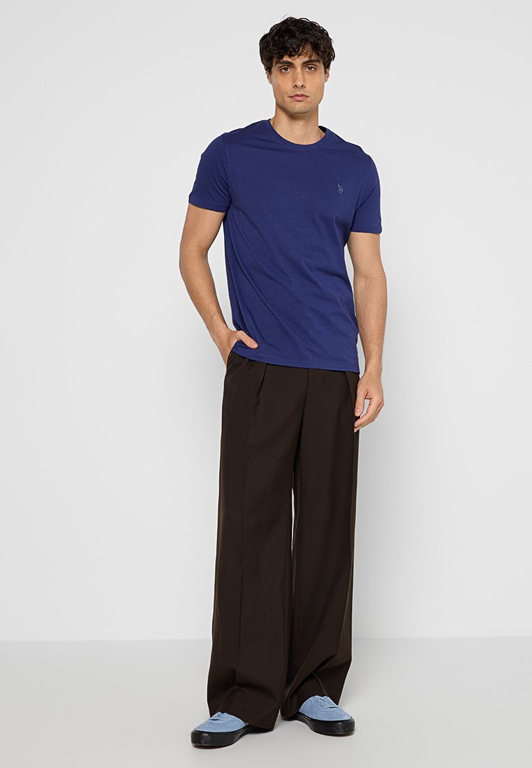 Dunkelblauses Baumwoll-T-Shirt mit kurzen Ärmeln, kombiniert mit weiten schwarzen Hosen und hellblauen Schuhen. Das Modell steht vor einem weißen Hintergrund.