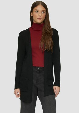 Frau mit langen braunen Haaren, die einen schwarzen Cardigan mit offenem Vorderteil, einen roten Rollkragenpullover und dunkle, hoch taillierte Jeans trägt, stehend vor einem grauen Hintergrund.