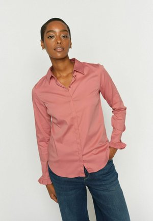 Žena s krátkymi vlasmi, oblečená v ružovej košeli na gombíky s volánmi na manžetách a modrých džínsoch, stojaca s jednou rukou v vrecku.