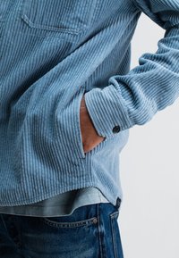 Person wearing blue corduroy jacket with hand in pocket, paired with blue jeans and a light blue shirt underneath.