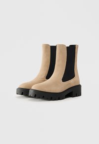 Beige semsket støvletter med svarte elastiske sidepaneler, rund tå og kraftig svart gummisåle med mønster.