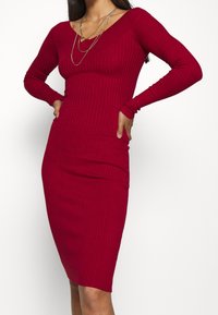 Robe rouge à côtes avec manches longues, décolleté en V et coupe ajustée. Fabriquée en tissu tricoté texturé, elle présente un design jusqu'au genou.