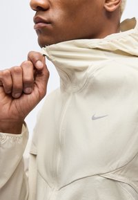 Personne à la peau brun clair et aux cheveux blonds courts remontant la fermeture éclair d'une veste Nike beige clair à col haut.