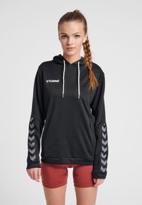 Felpa nera con cappuccio e cordini bianchi, caratterizzata da un tessuto strutturato e accenti a zigzag grigi sulle maniche, abbinata a pantaloni corti rossi.