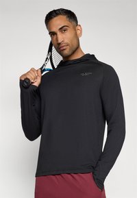 Melnā garo piedurkņu sporta krekls ar kapuci, logotipu uz krūtīm un gludu tekstūru, to valkā modelis, kurš tur tenisa raketi.