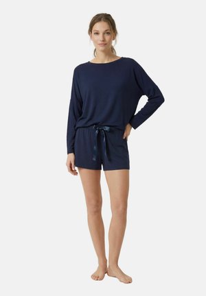 Femme debout pieds nus portant un haut à manches longues bleu marine et un short assorti avec un ruban noué à la taille sur fond blanc.