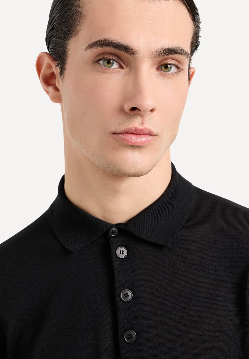 Polo shirt nera realizzata in tessuto liscio, con colletto classico e patta con quattro bottoni di colore scuro. Vista ravvicinata del collo.
