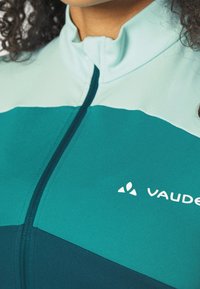 Teal zipjacka med ljusare teal-accenter och en mörkblå sektion. Har en upphöjd krage och en vit logotyp på bröstet. Slät tygtextur.