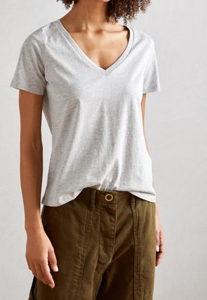 Person trägt ein hellgraues V-Ausschnitt-T-Shirt mit kurzen Ärmeln, das leicht in eine hoch taillierte olivgrüne Hose mit Knopfverschluss gesteckt ist.