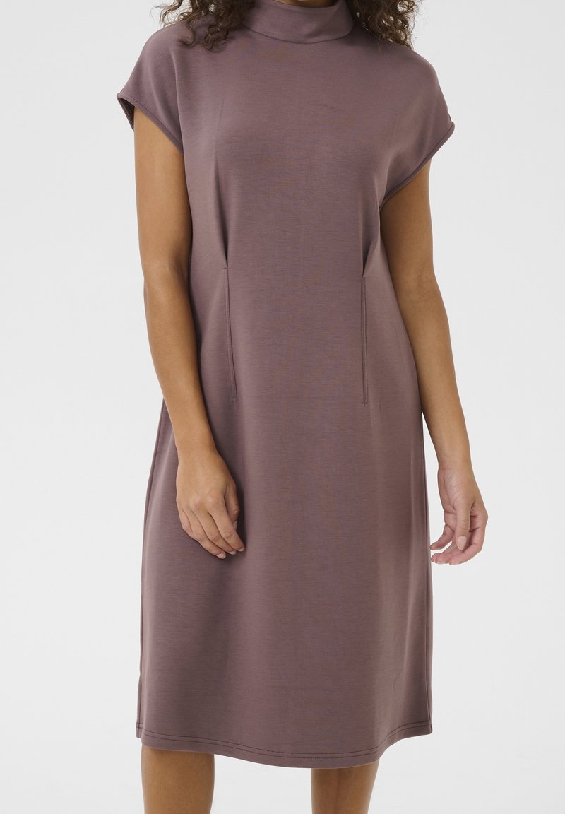 Femme portant une robe mauve sans manches longueur genou avec un col montant, debout devant un fond gris clair uni.
