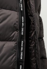 Gros plan d'une fermeture éclair de veste noire avec un texte blanc sur un ruban en tissu indiquant « KINGS WILL DREAM » à plusieurs reprises.