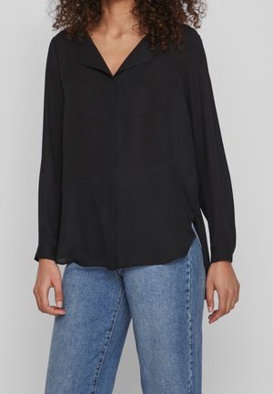 Vrouw draagt een losse zwarte blouse met lange mouwen en een V-hals kraag en lichtblauwe spijkerbroek, staand tegen een effen achtergrond.