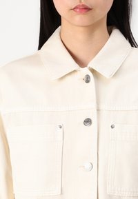 Chaqueta de mezclilla beige con un cuello clásico, botones a presión plateados, dos bolsillos en el pecho y un acabado de tela texturizada.