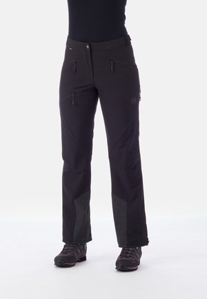 Zwarte outdoorbroek met ritszakken gedragen met zwarte wandelschoenen bij een persoon die tegen een witte achtergrond staat.