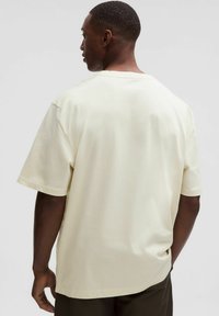 Tričko s krátkym rukávom v krémovej farbe, vo voľnom strihu. Má okrúhly výstrih a hladkú textúru, zobrazené zozadu.