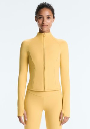 Nő szűk, sárga cipzáras dzsekit és hozzáillő leggingset visel, egyszínű világos háttér előtt áll.