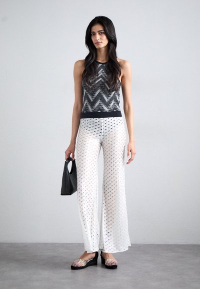 Top crop cu model alb și negru, pantaloni albi largi, transparenți, cu o textură asemănătoare unei plase, geantă neagră și pantofi cu toc multicolori.