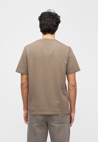 Brązowa koszulka z krótkim rękawem wykonana z miękkiego materiału o luźnym kroju. Posiada okrągły dekolt i gładką fakturę, noszona z jasnymi spodniami.