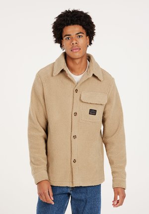 Beige fleecejack voor dames met knoopsluiting aan de voorkant, kraag, één borstzak en een merkembleem. De stof heeft een gestructureerde, gezellige uitstraling.