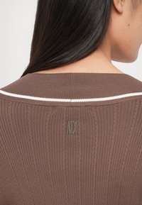 Pull en maille côtelée marron avec une bande blanche le long de l'encolure, arborant un petit logo gris à l'arrière. Texture douce et coupe ajustée.