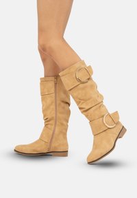 Botas altas de ante en tono marrón claro con cremallera lateral, diseño arrugado y dos tiras decorativas con hebillas circulares doradas.