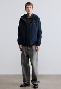 Ελαφρύ σακάκι σε ναυτικό μπλε με κουκούλα και λάστιχο στα μανίκια, φορεμένο πάνω από σκούρο γκρι πόλο. Συνδυάζεται με χαλαρά γκρι τζιν και μαύρα παπούτσια.