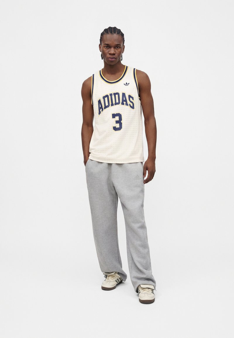 Young man standing wearing a white Adidas sports tank top, grey sweatpants, and light-colored sneakers on a plain white background.