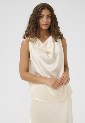 Lokkisprendiga pruunide juustega naine, kes kannab varrukateta kreemjat satiinist topi ja sobivat seelikut, seisab ühevärvse heleda tausta ees.