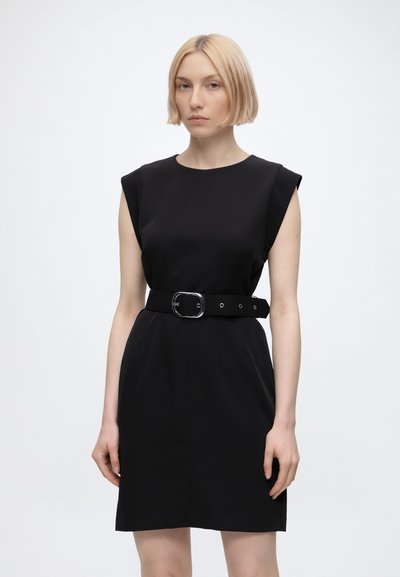 Femme aux cheveux blonds courts portant une robe noire sans manches avec une large ceinture et une boucle argentée, debout devant un fond blanc uni.