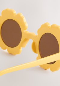 Rumene sončna očala v obliki cvetov, z rjavimi lečami. Dizajn vključuje teksturiran okvir in široke, elegantne stranice.