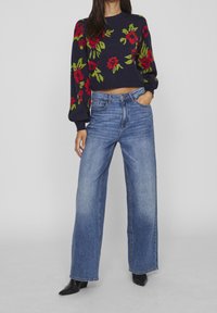 Pull marine avec un motif floral rouge et vert, associé à un jean denim bleu à jambes larges. Texture lisse avec un design raccourci. Bottines noires.