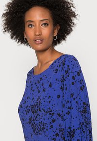 Camisola azul com um corte descontraído, apresentando um padrão de respingos pretos, gola redonda e mangas compridas. Textura suave. Brincos simples em forma de argola.