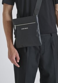 Borsa a tracolla nera con texture, dotata di zip argentata, con una tasca frontale e nome del brand in rilievo, indossata sopra una maglietta nera.