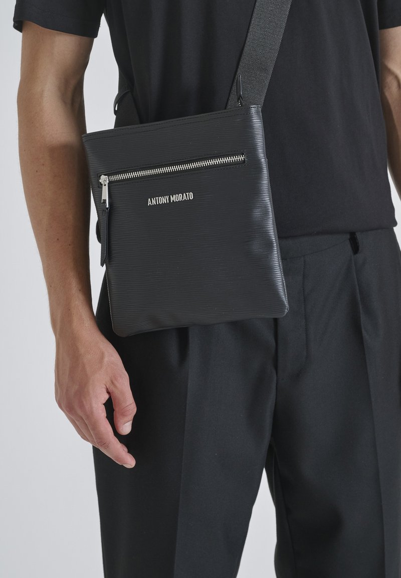 Borsa a tracolla nera con texture, dotata di zip argentata, con una tasca frontale e nome del brand in rilievo, indossata sopra una maglietta nera.