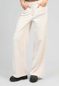 Pantalons en velours côtelé beige clair, à jambes larges, avec poches avant, fermeture à bouton et légère texture à nervures verticales. Portés avec des bottes compensées noires.