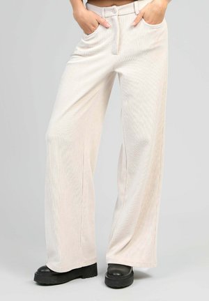 Pantalons en velours côtelé beige clair, à jambes larges, avec poches avant, fermeture à bouton et légère texture à nervures verticales. Portés avec des bottes compensées noires.