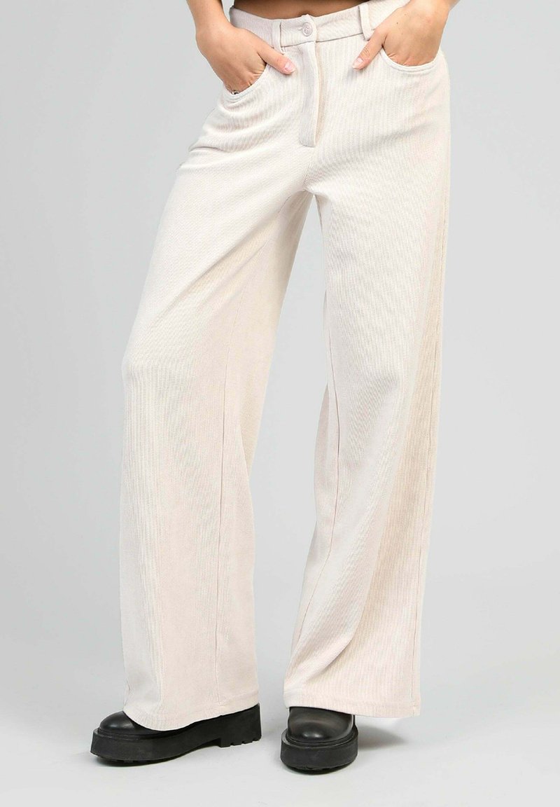 Pantalons en velours côtelé beige clair, à jambes larges, avec poches avant, fermeture à bouton et légère texture à nervures verticales. Portés avec des bottes compensées noires.